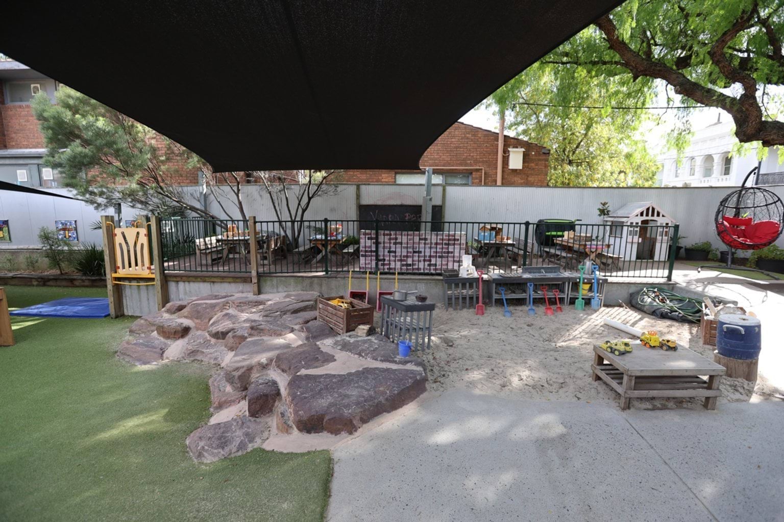 Playground overview showing artificial turf, rock formations, sand area and timber climbing equipment under shade sails.