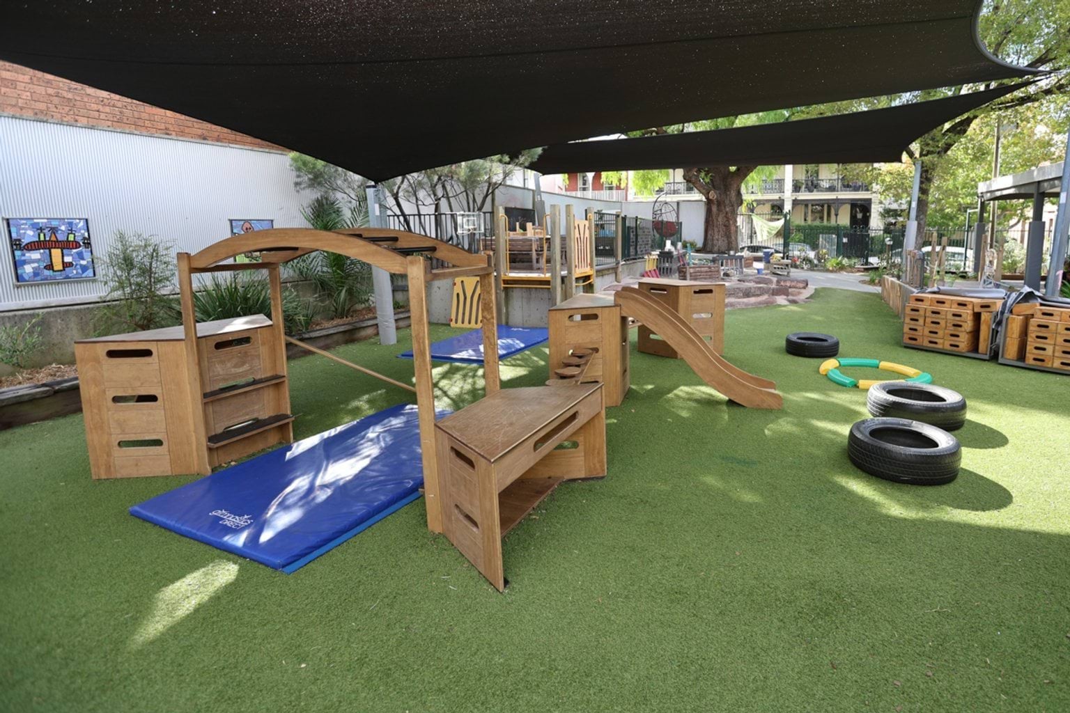 Wide view of timber climbing structures, slides and balance elements on artificial grass in a shaded playground.