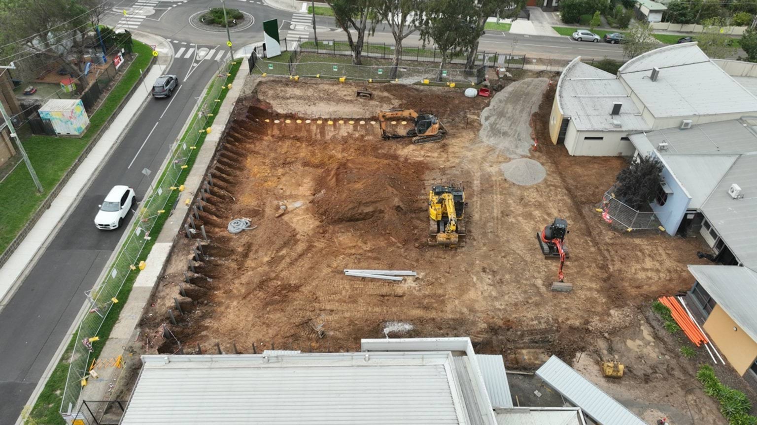 Aerial view of a school construction site with excavated ground and earthmoving machinery beside a road and school buildings.