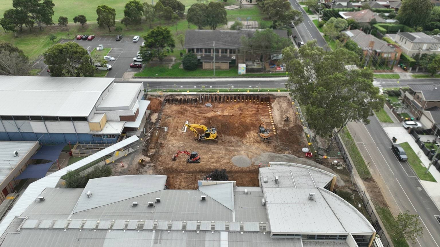 Wide aerial view of a school campus with a central excavation site, construction machinery and nearby streets and trees.