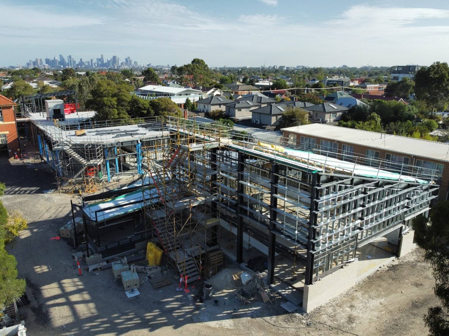 Aerial view of a steel‑framed school building under construction, with scaffolding, materials on site and nearby homes in the background.