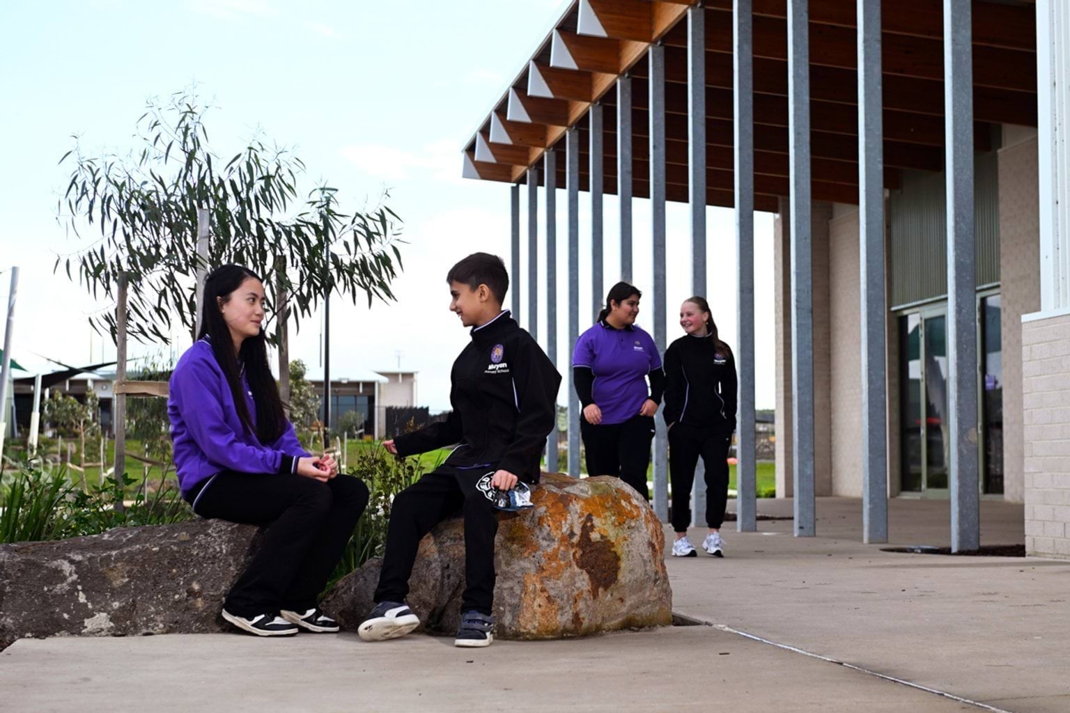 4 kids in purple and black school uniforms are outside their new school. 2 are sitting on rocks bordering a garden and 2 are in the background walking.