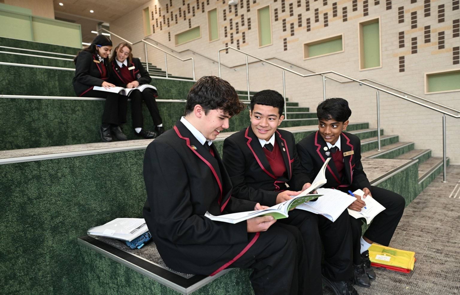 5 kids in maroon and black school uniform are sitting on carpeted stairs and looking at their school books together. 2 girls are at the top and 3 boys are seated closer to the front.