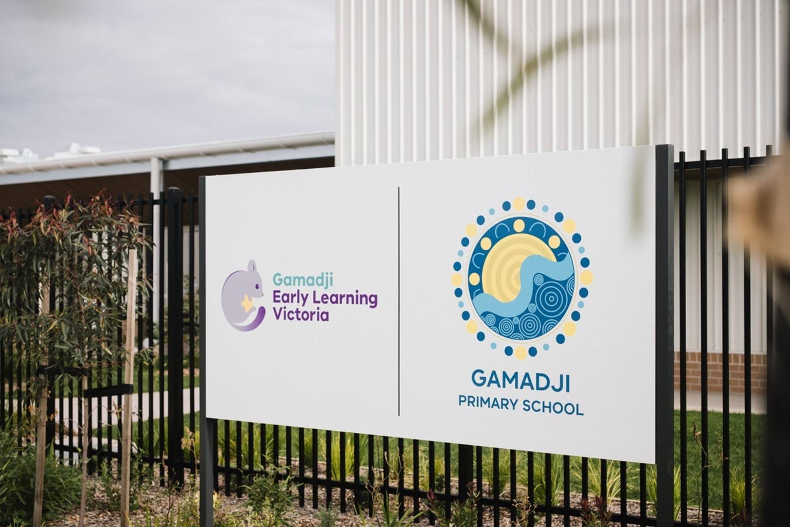 Black fence with sign of two logos: Gamadji Early Learning Victoria and logo of purple possum; and Gamadji Primary School with logo of blue and yellow indigenous style sun and ocean