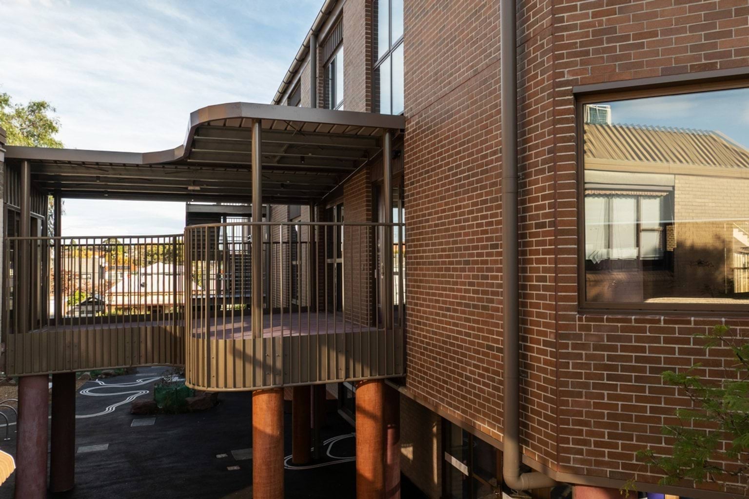 Brown brick building with curved, raised walkway with shelter. We can see paved concrete area below and the building's windows