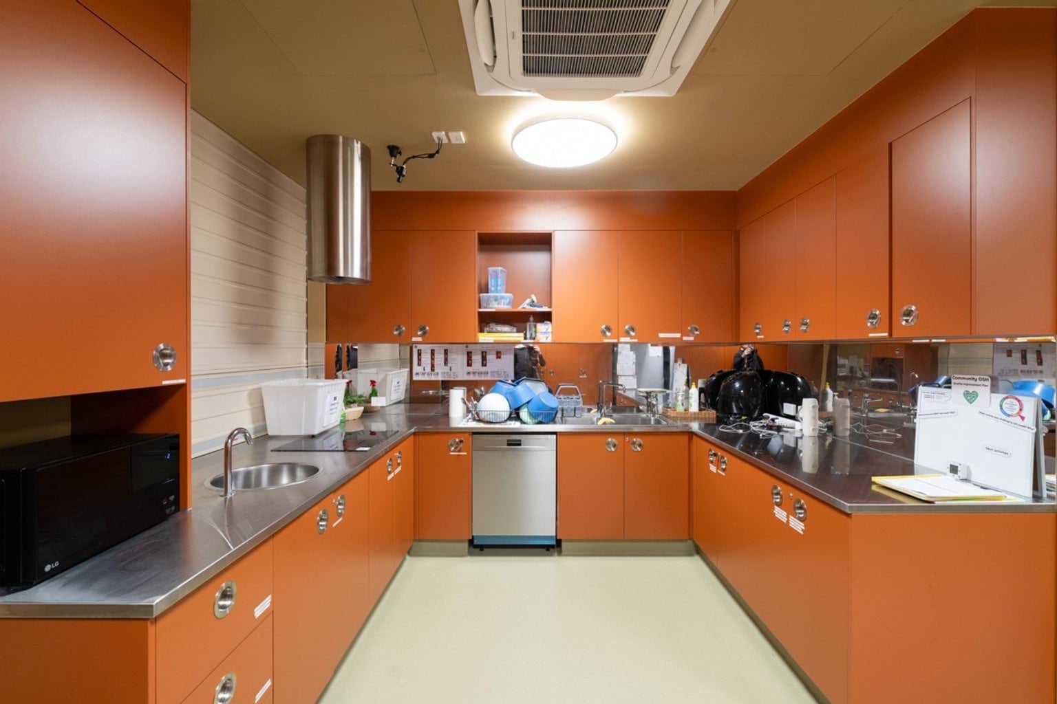 Open kitchen space with orange cabinetry and metal benchtops. We can see a sink and induction stovetop on the left hand side and a dishwasher and drying area in the centre.