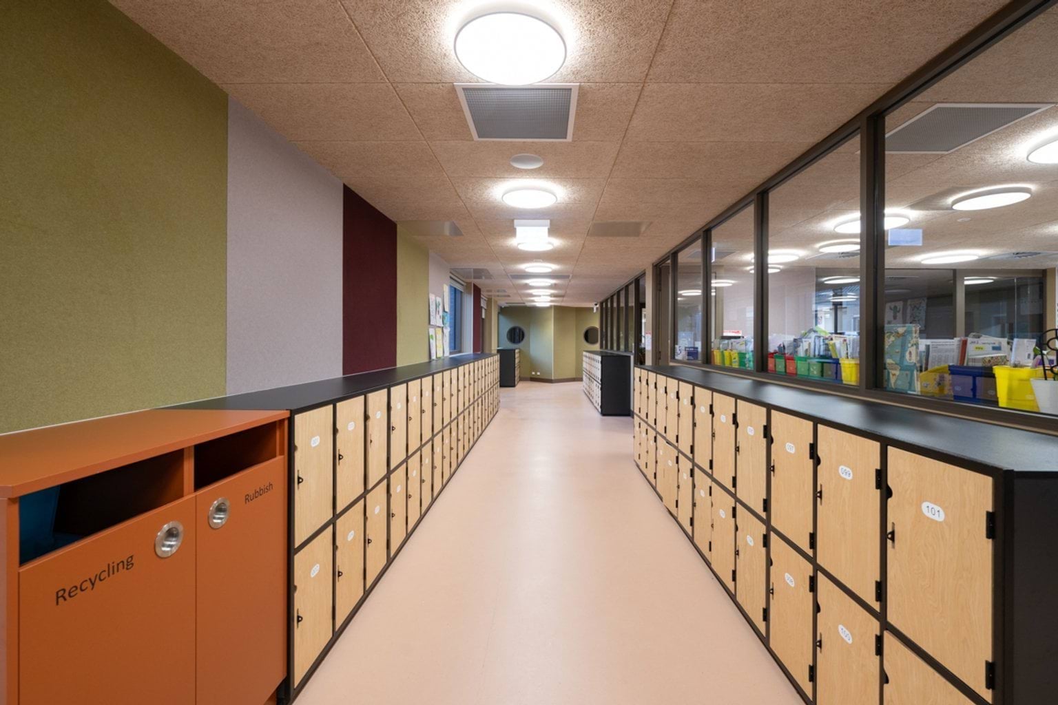 Hallway with lockers on each side, orange recycling and rubbish bins, padded multicolour felt walls and a window which sees into a classroom on the right hand side.