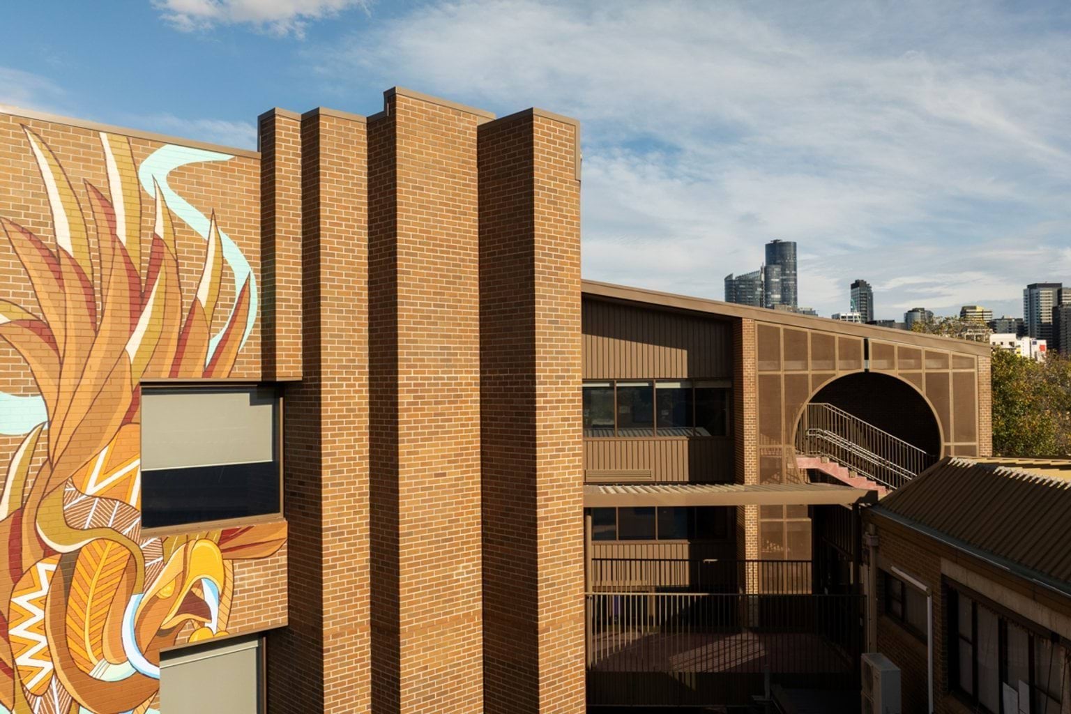 Brown brick buildings with indigenous artwork of Bunjil on the wall and a partly cloudy city skyline in the background
