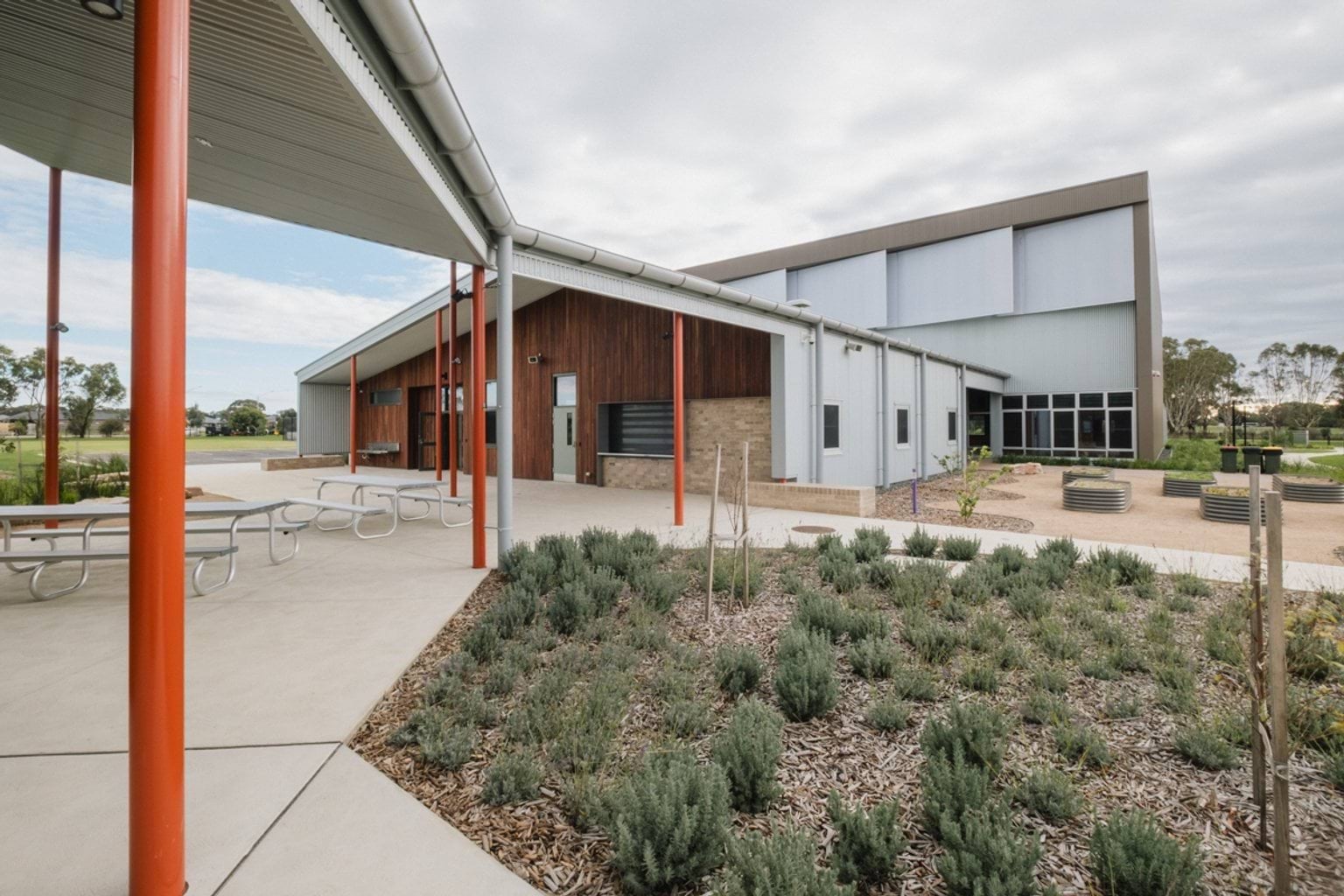 Outdoor covered area with garden beds. We can see long tables and benches under the covered area attached to a building. There's vegetable garden boxes next to the building