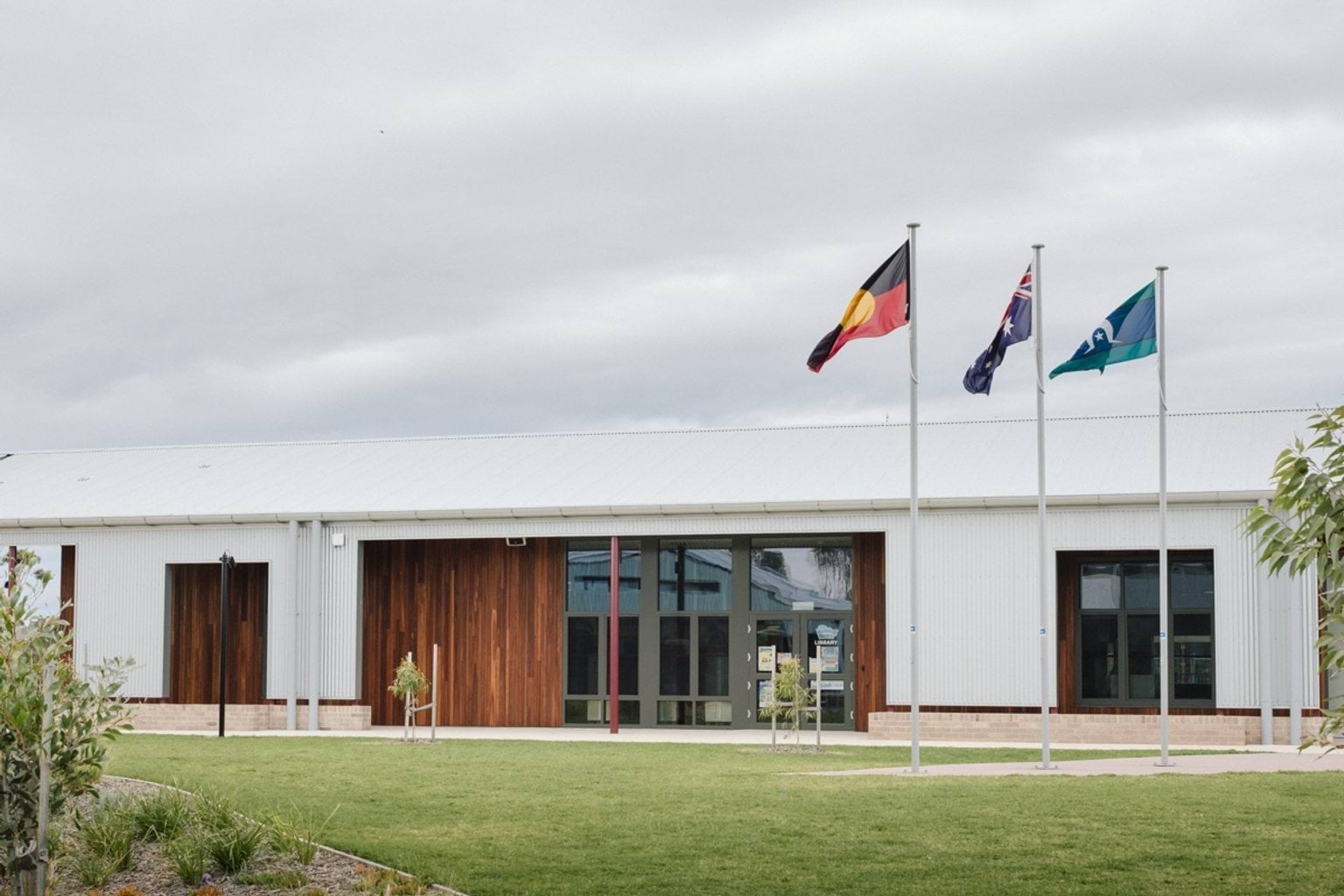 Single storey white building with wooden panelling and windows. In the foreground we can see Indigenous, a Torres-Strait and Australian flagpoles and grass lawn with garden beds