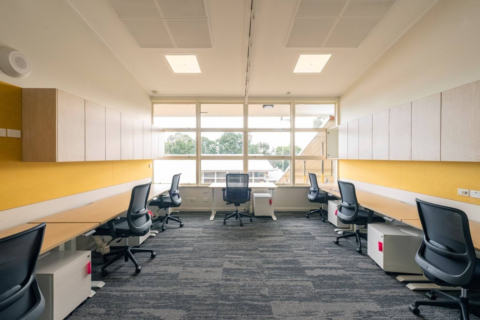 A bright office space with rows of desks and office chairs along both sides of the room and large windows at the rear.