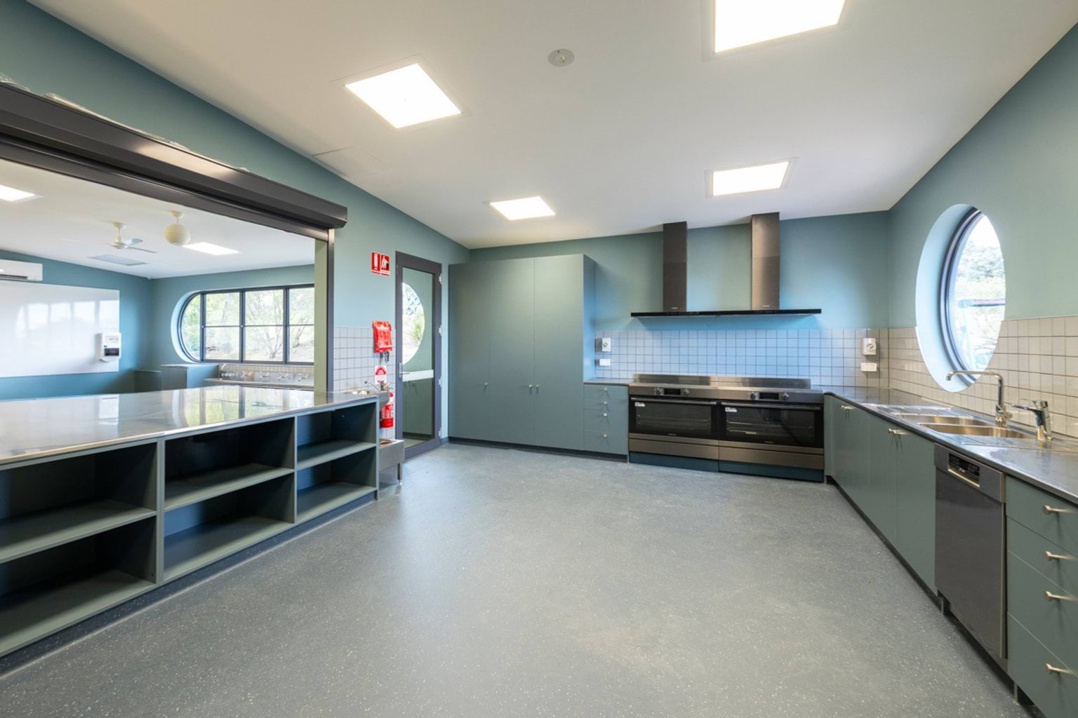 A brightly-lit kitchen with shelves, stoves, ovens, rangehoods and a dishwasher.