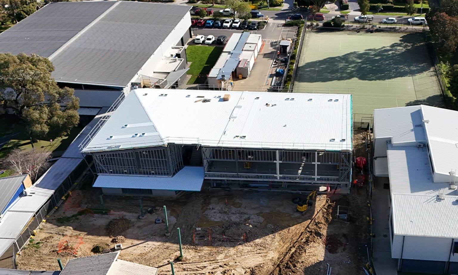 Aerial view of a large building under construction, with exposed ground and construction equipment in the foreground and completed buildings surrounding the site.