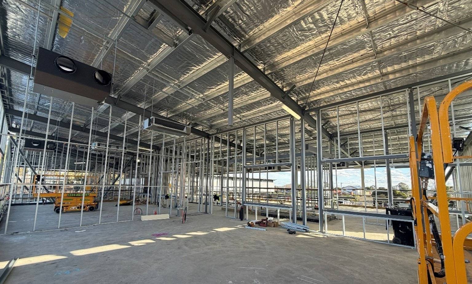 Interior view of a building under construction, showing exposed steel framing, insulation-lined roofing, and open floor space with construction equipment and materials visible throughout.