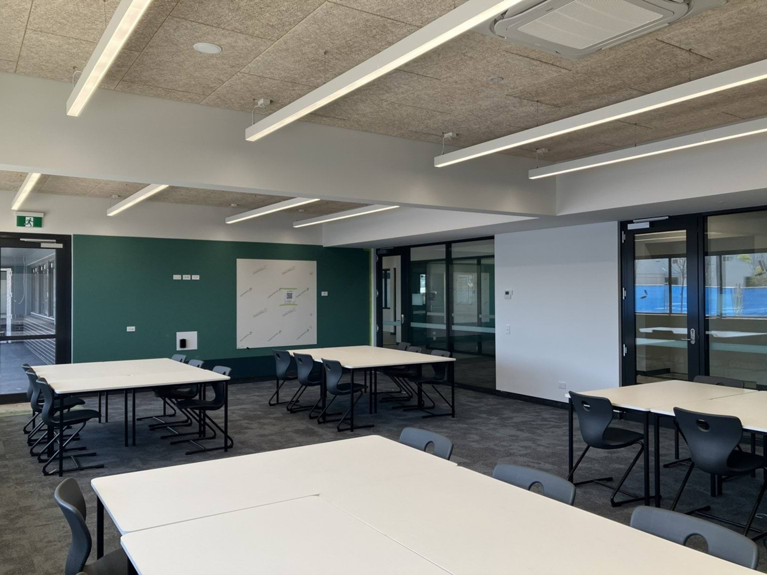 A classroom with tables and chairs. The room includes large windows and overhead lighting.