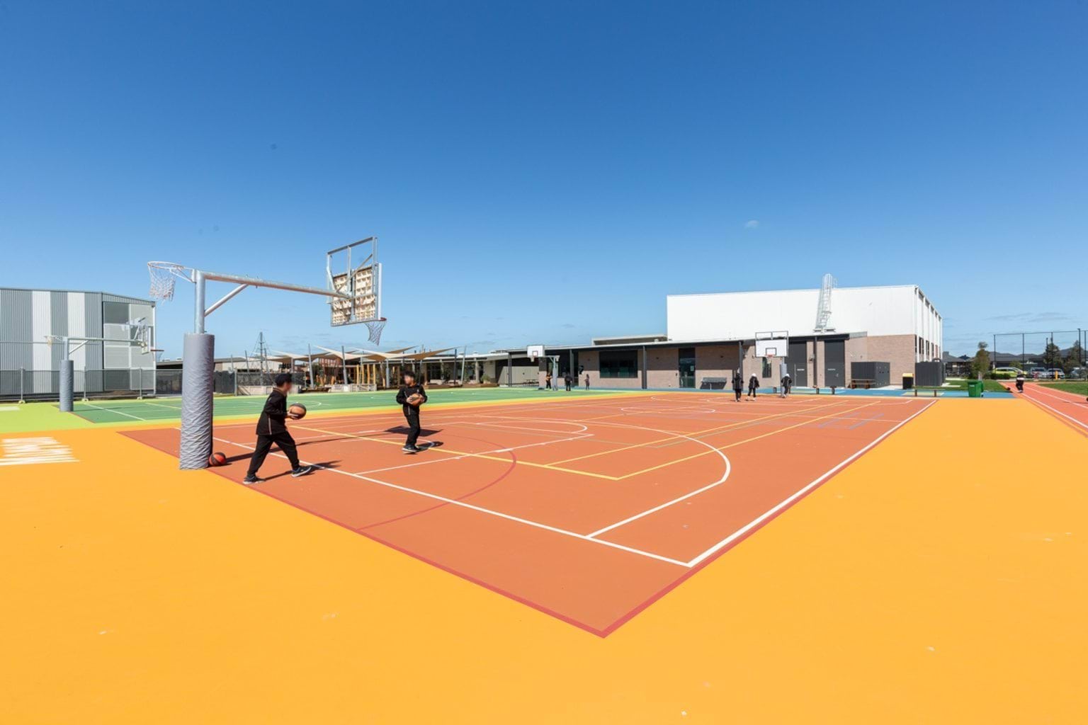Multi-coloured outdoor court areas with basketball and netball rings. We can see a school building in the background and two students playing on the court.