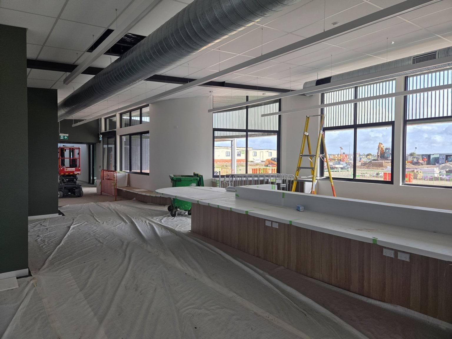 Foyer area under construction featuring a long built-in bench or counter beneath large windows. Protective floor coverings, a ladder, exposed ceiling services and unfinished ceiling panels and large windows are visible.