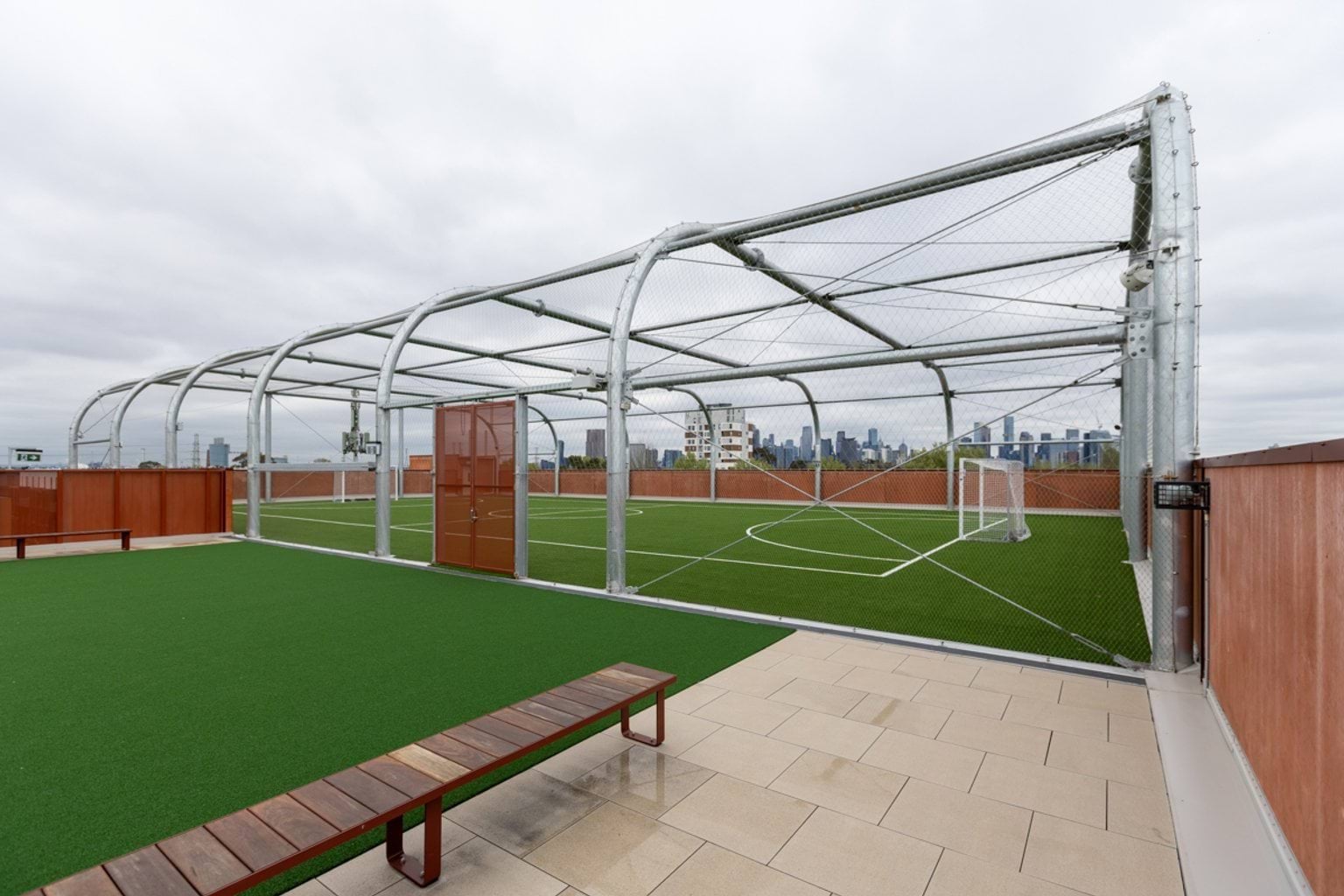 A rooftop soccer field in a cage. There is a tiled paved area and a wooden bench seat.