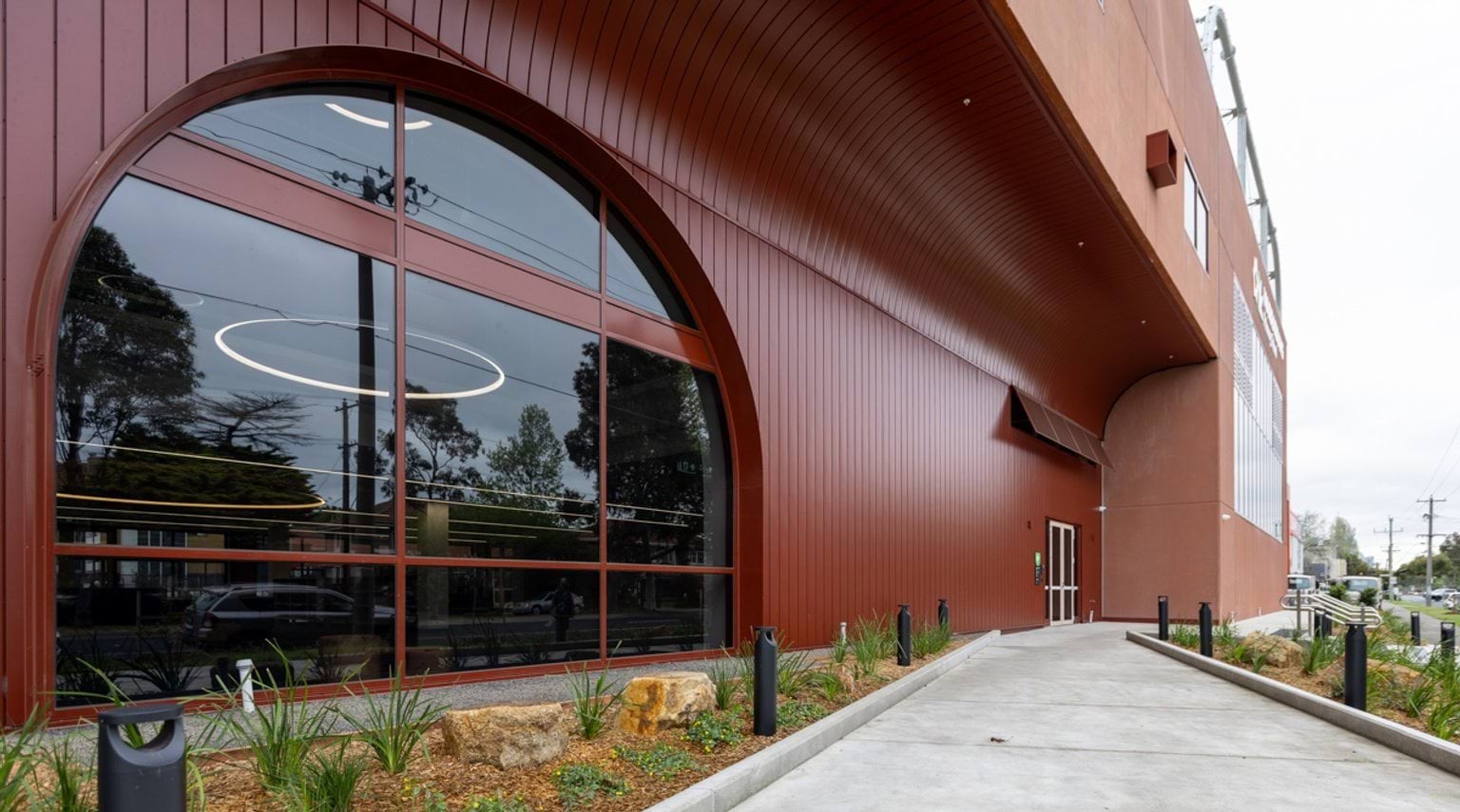 Paved pathway with landscaped garden beds on either side leading to a multi-level red/orange building with a large arched window.