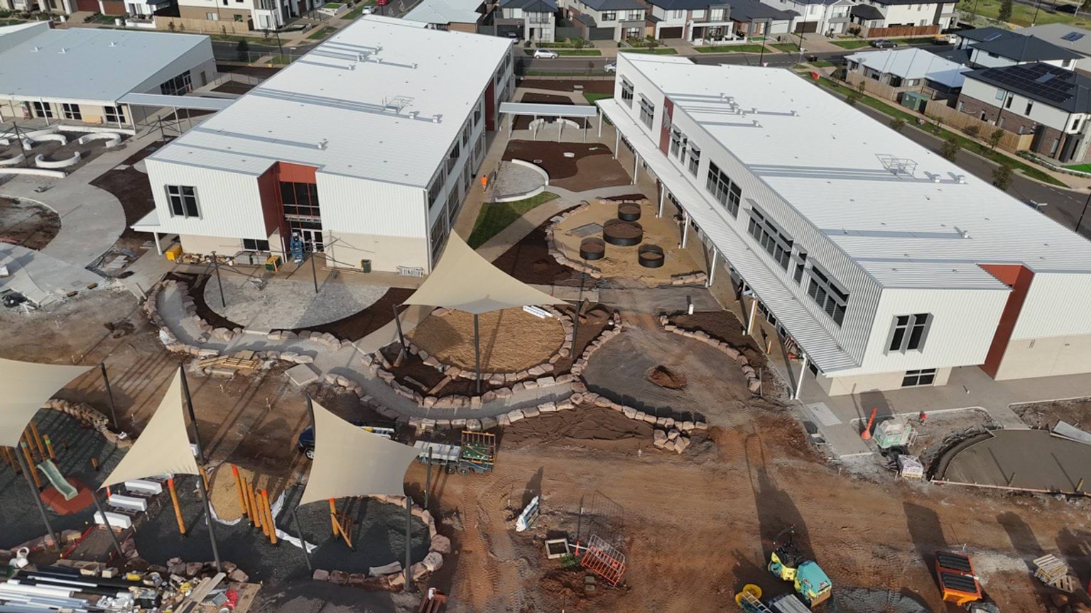 Aerial view of newly constructed school buildings surrounding a central landscaped courtyard with outdoor learning and play areas that are still being completed.